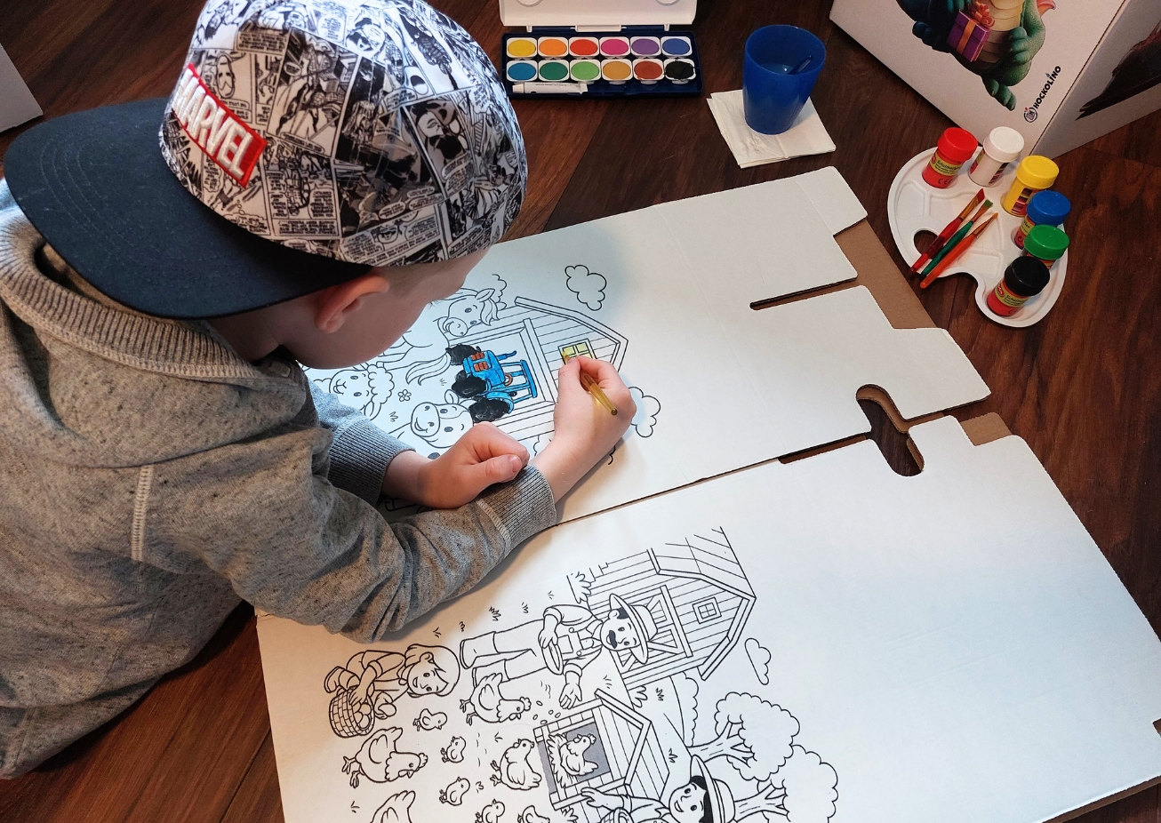 Kind malt mit Buntstiften auf einem Hockolino mit einer Bauernhofszene auf dem Boden.